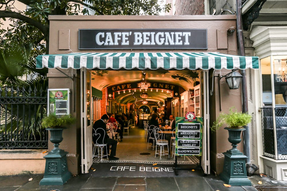 The front of Cafe Beignet, a famous cafe in New Orleans, with a striped green and white canopy and black tile in front of a timeless-looking restaurant.