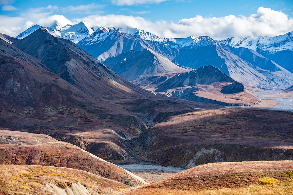 Several snowcapped mountains and rolling hills against a blue, cloudy sky in Denali National Park, one of the best places to visit in the USA in July.