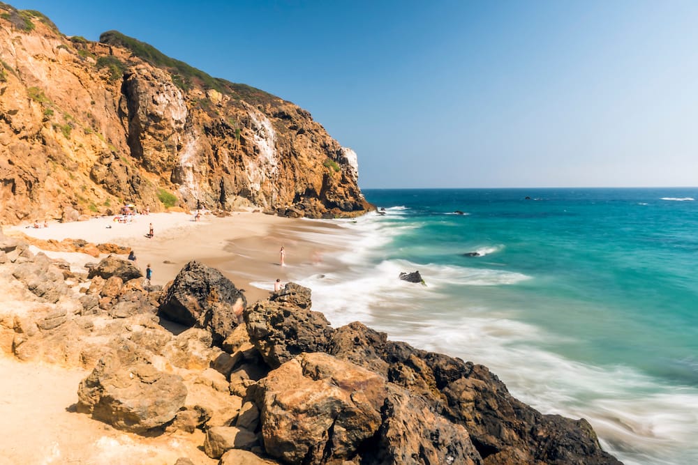 Turquoise waves lapping onto a golden sand beach with golden rocks reaching out onto the sandy beach.