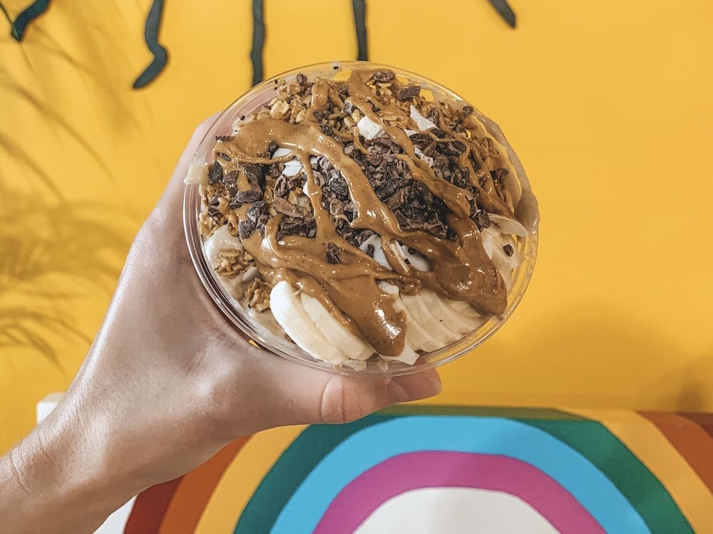 A bowl with bananas, chocolate, peanut butter, and other toppings packed on top in front of a yellow wall with a rainbow painted on it.