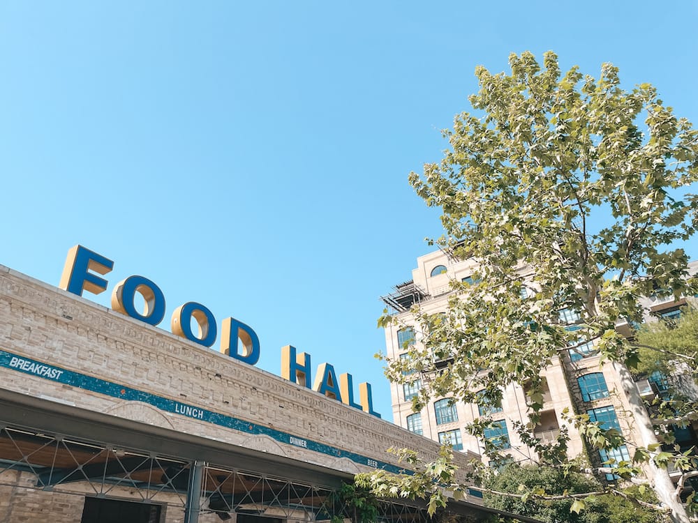The blue sign for the food hall at the Pearl in San Antonio, with a tree in the foreground covering Hotel Emma in the background.