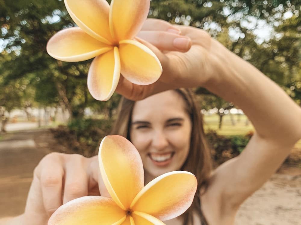 A girl holding two yellow and pink pulmeria flowers toward the camera in a park in Waikiki, Hawaii