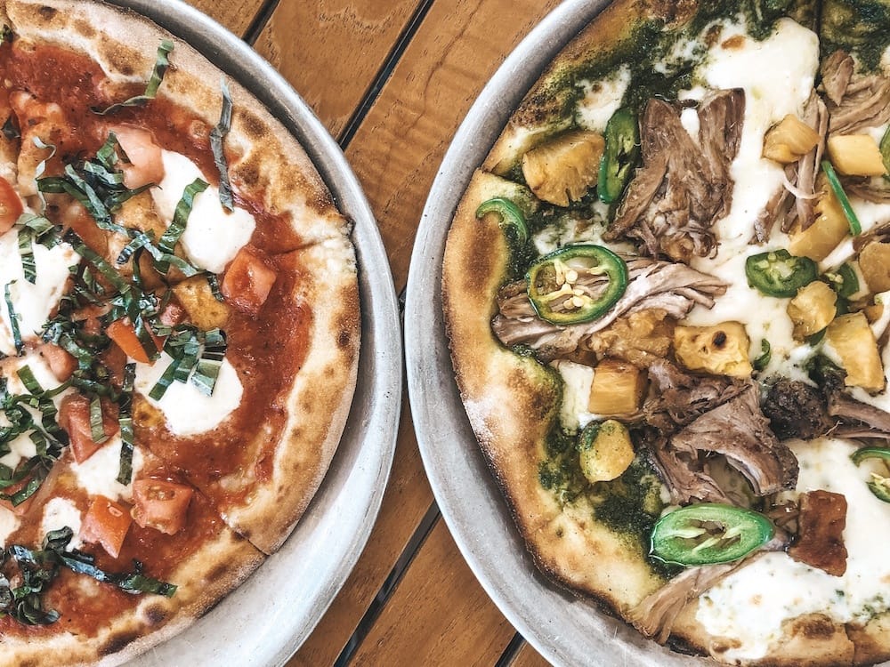 Two pizzas sitting on a wooden table in Waikiki, one with red sauce and mozzarella and another with Kalua pork, jalapenos, and pineapple.