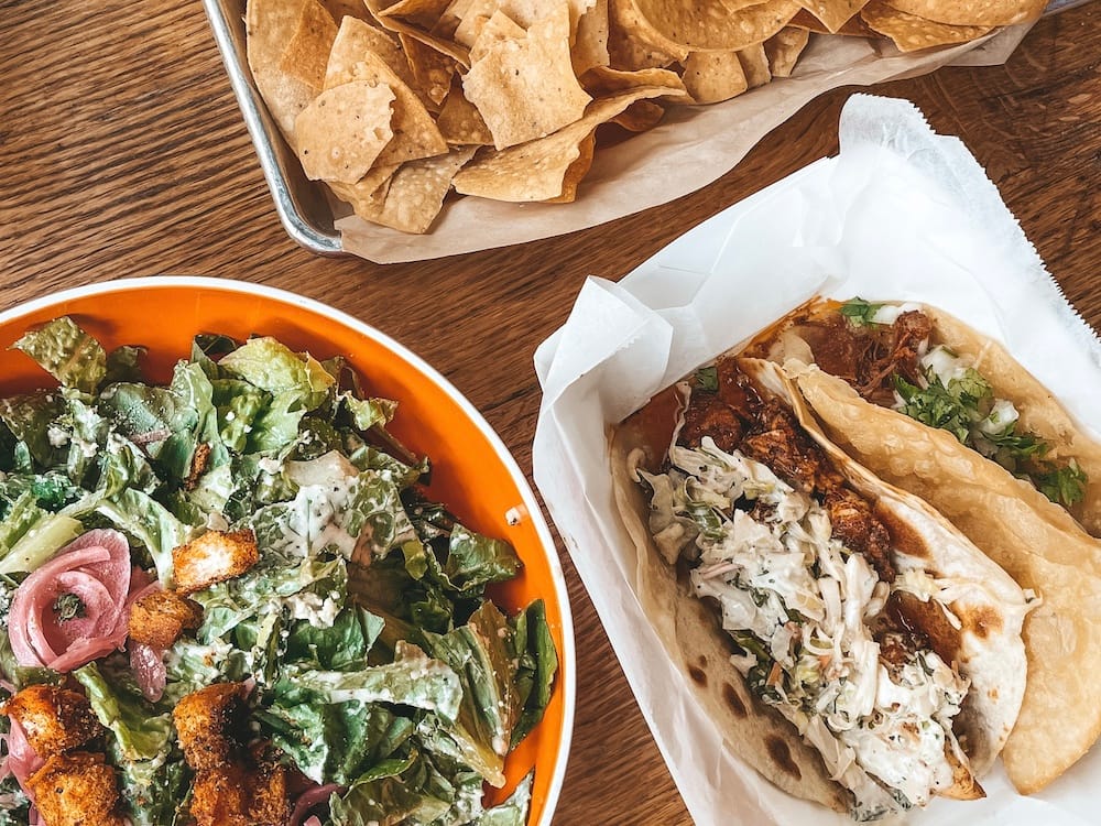 A ceasar salad, two tacos, and tortilla chips sitting on a table.