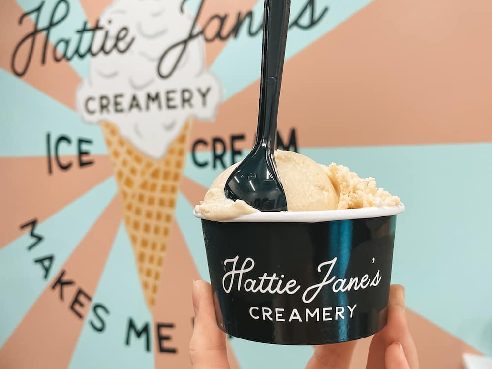 A scoop of ice cream in a cup from Hattie Jane's Creamery being held in front of a colorful pink and blue mural inside the scoop shop.