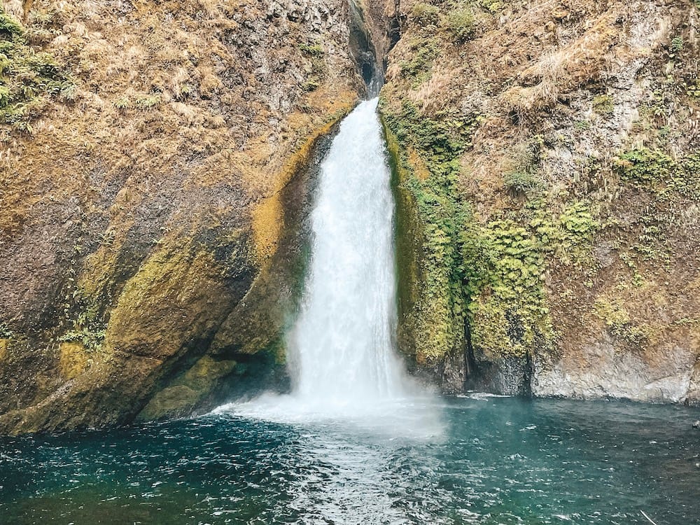 A waterfall cascading off a mossy, rocky cliff into a pool of bluish-green water.