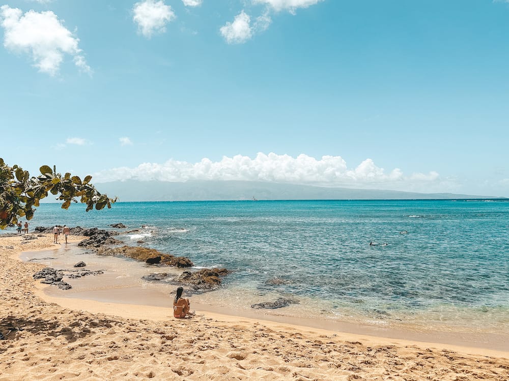 The blue ocean water at Napili Bay in Maui, with a girl sitting in the golden sand and looking out at the water.