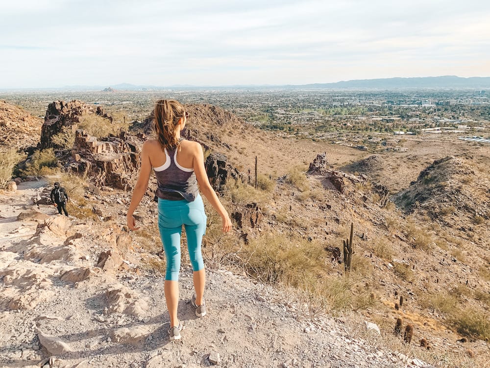 A woman with a brown ponytail, grey tank top, and green leggings standing on a hiking trail on a mountain in Scottsdale overlooking the desert