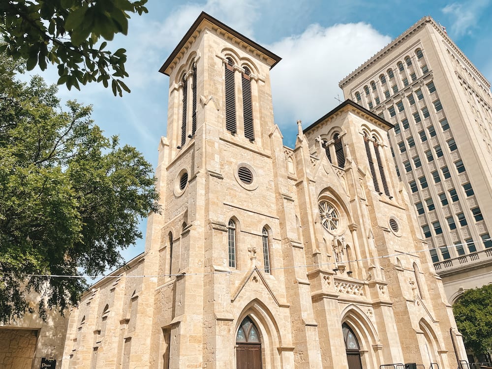 A tall, historic church with ornate details built from tan bricks stands against a blue sky with a green tree in the background in San Antonio. The San Fernando Cathedral is one of the best things to do during a weekend in San Antonio