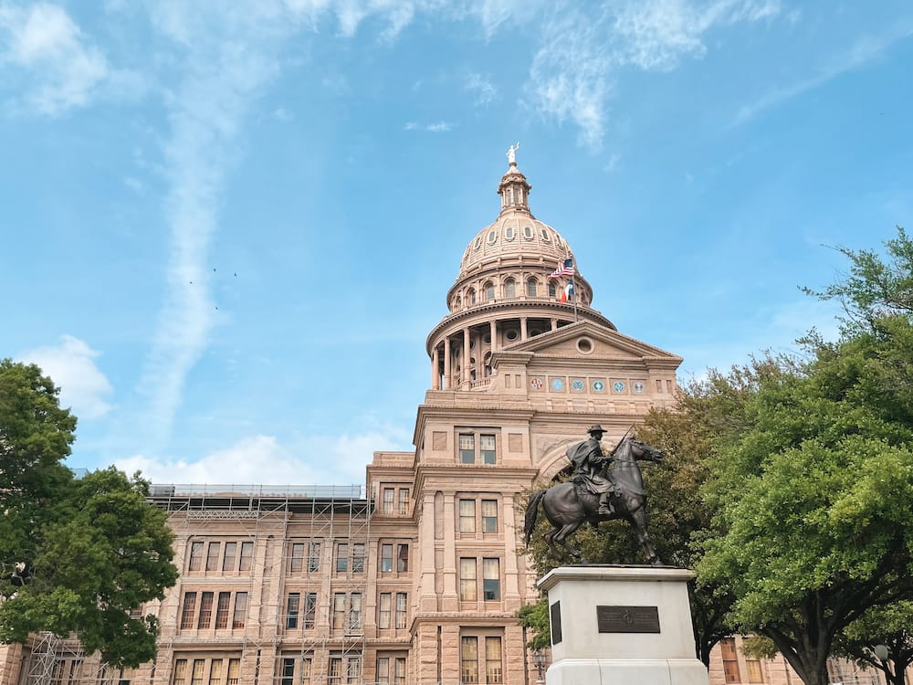 The Texas State Capitol building against a clear blue sky. There is an iron statue of a man riding a horse in front of the building.