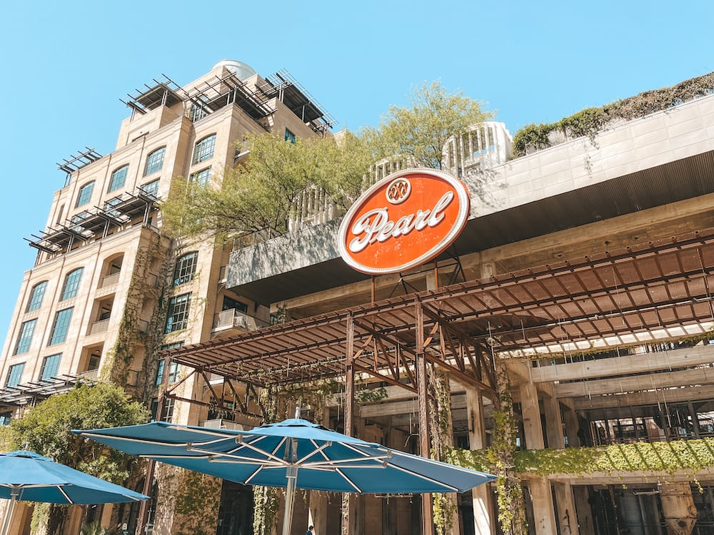 The red sign for The Pearl, a shopping and dining district in San Antonio. The sign is fixed on a historic building that features greenery trailing up and down the sides, and there are blue umbrellas in front of it.