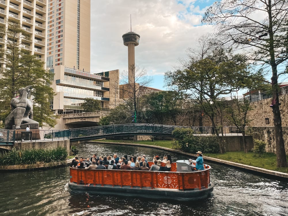 A boat cruising down the San Antonio River Walk in front of the Tower of the Americas, one of the tallest observations towers in the United States.