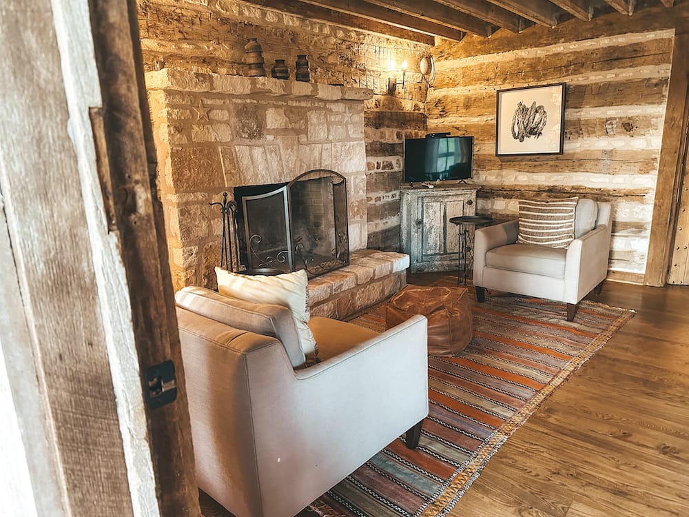 A cozy living room in a log cabin with two chairs, a TV, a stone fireplace, and a colorful striped rug.