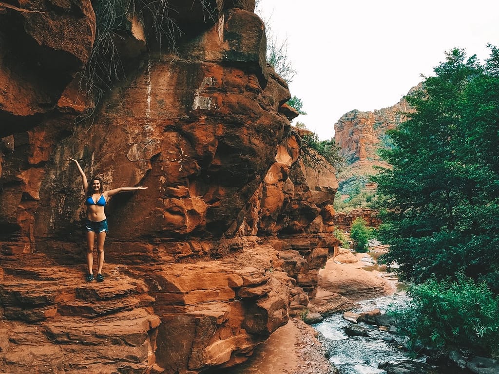 A woman wearing a blue bikini top and denim shorts standing on stop of a red rock overlooking Slide Rock State Park.