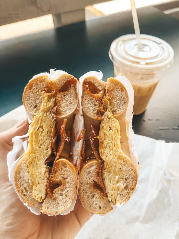 A bagel split it half, revealing eggs and bacon inside with an iced coffee in the background.