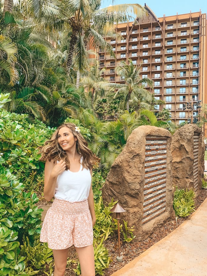 A girl in a white tank top and pint shorts standing in front of a lush green bush and several palm trees with a resort in the backgorund