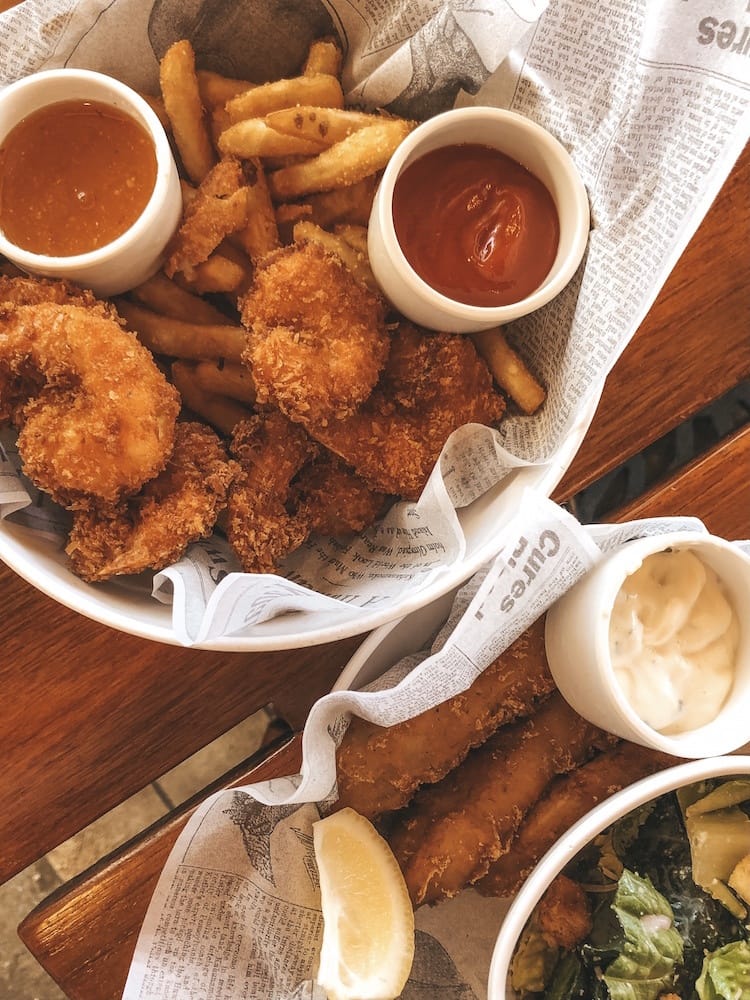 A basket of coconut shrimp and fried and another basket with fish and chips sitting on a wooden table at Duke's Waikiki.