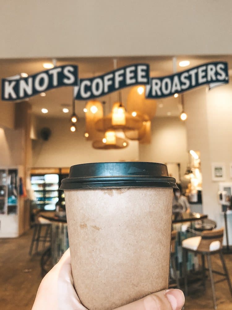A brown, disposable cup of coffee with a black plastic lid being held in front of the sign for Knots Coffee Roasters, one of the best coffee shops in Oahu.