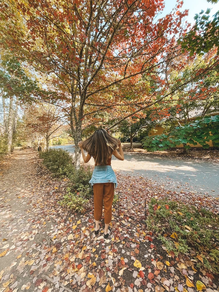 A woman with long brown hair wearing an orange jumper standing on front of a tree with red leaves during the fall.