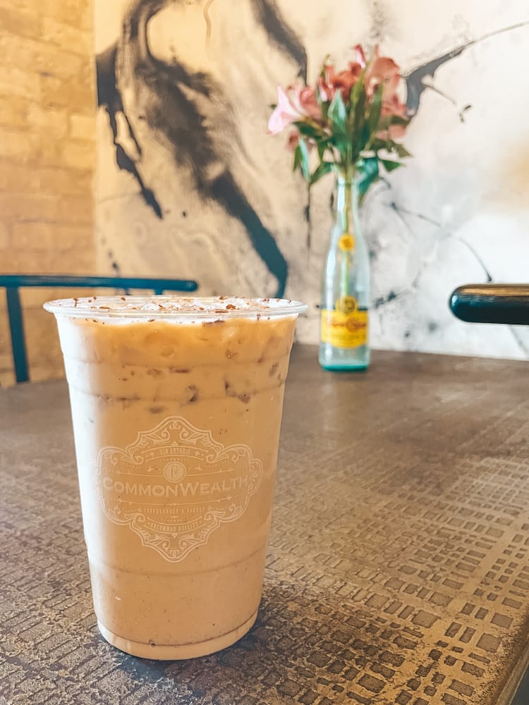 An iced coffee sitting on a wooden table with a vase of flowers in a Topo Chico bottle in the background