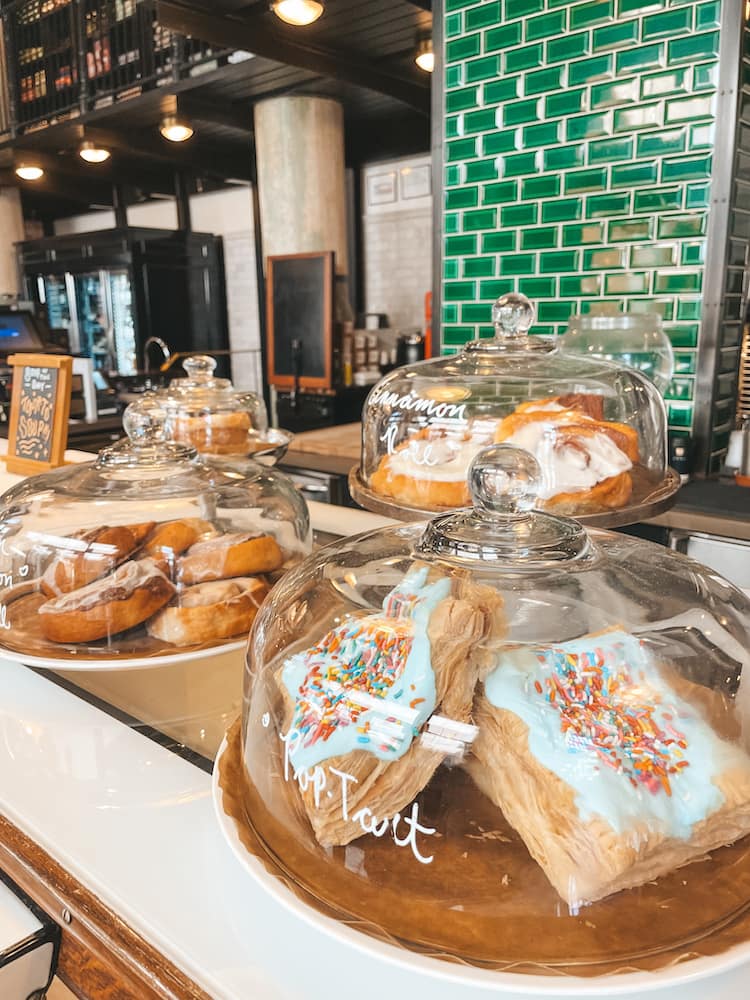 Several pastries, like pop-tarts and cinnamon rolls, are sitting in cake display cases in a coffee shop. The pastries are sitting in front of a green tiled wall.