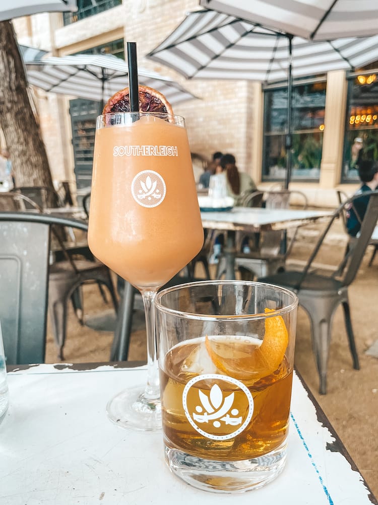 Two drinks sitting on a metal table with black and white umbrellas and a brick building in the background. One is a pink slushy in a wine glass, and the other is a bourbon drink with an orange peel used as a garnish.