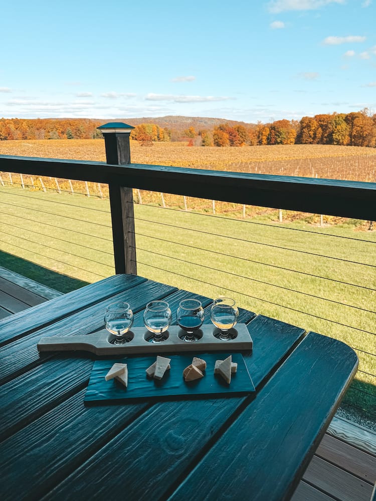 A flight of four wines and four cheeses sitting on a wooden table at one of the best wineries in the Finger Lakes, Stever Hill Vineyards, with a grassy field, vineyard, and fall foliage in the background.