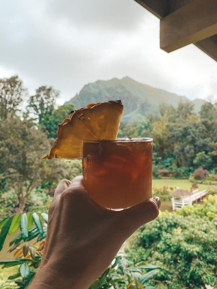 A tropical cocktail with a pineapple in it in front of a lush, green, tropical mountain range and landscape.