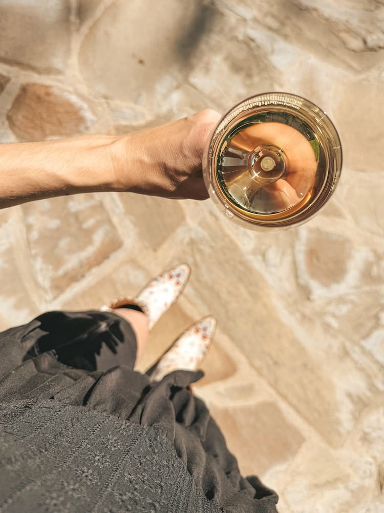 A glass of white wine held above a flagstone walkway, being held by a woman in a black dress and white cowboy boots.