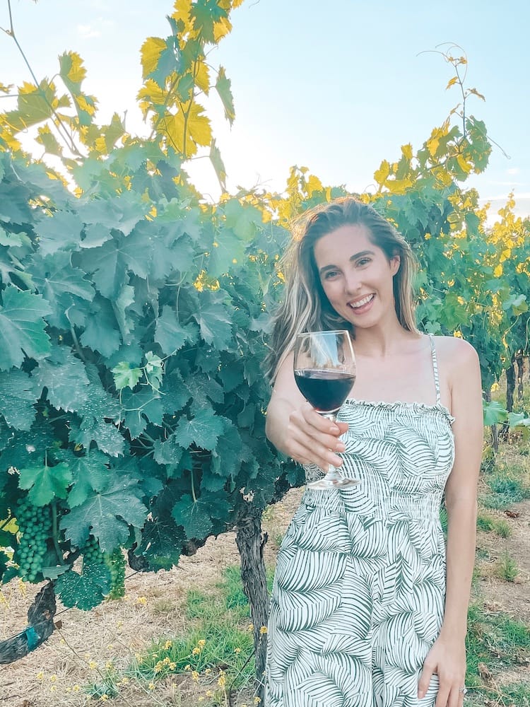 A woman wearing a green and white jumpsuit holding out a glass of red wine in front of a vineyard at one of the best wineries in Temecula.