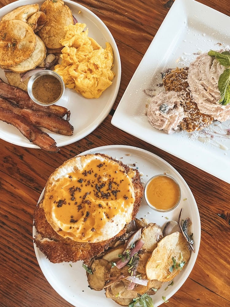 A breakfast spread of fried chicken, French toast, and eggs, bacon, and potatoes sitting on a wooden table.