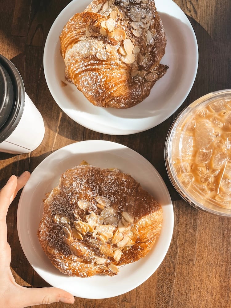 Two almond croissants sitting on a wooden table with a hot coffee and an iced coffee.