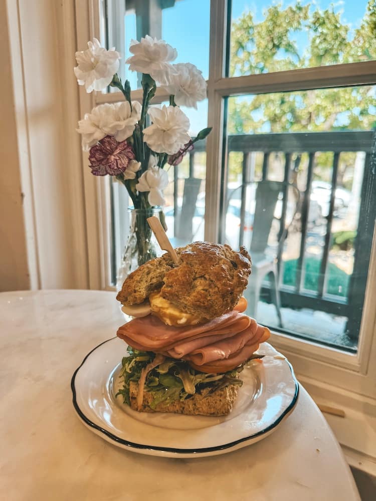A sandwich made from a scone sitting on the table in front of a vase of flowers and a window.