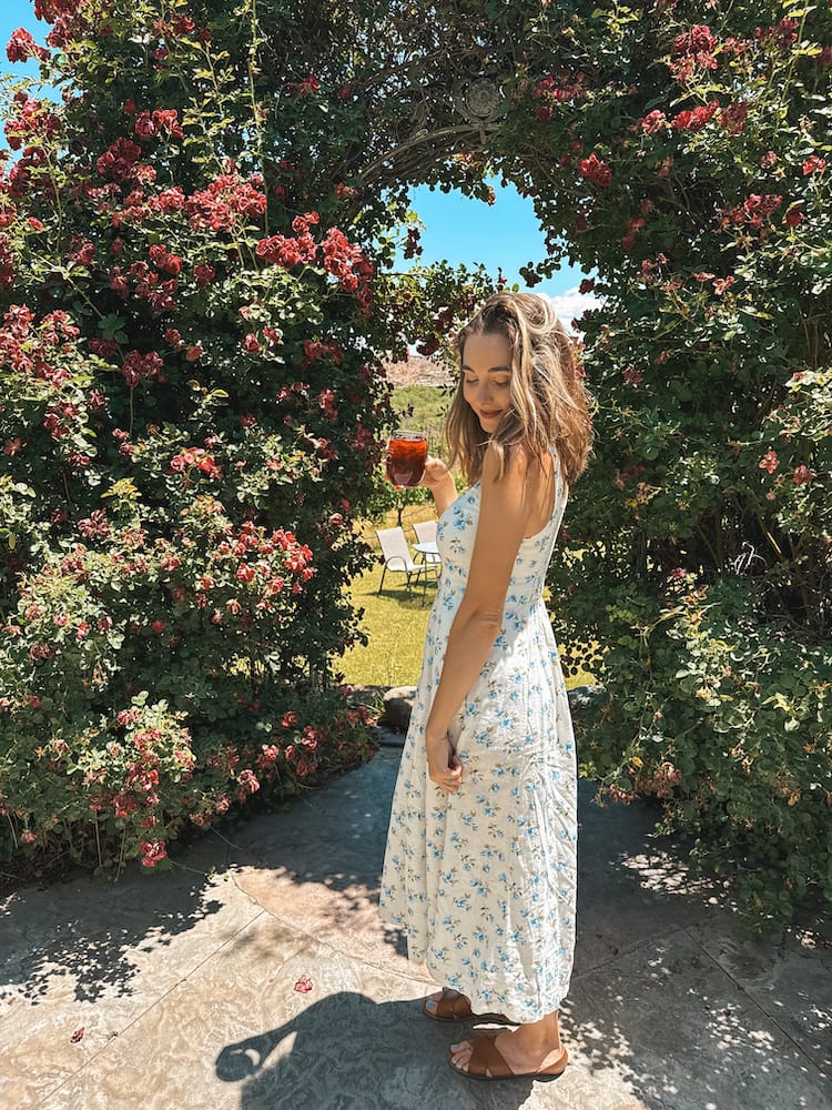 A woman in a blue and while floral sundress standing in front of an arch of bougainvilleas holding a glass of red wine at Alcantara Vineyards, one of the best wineries in Sedona.