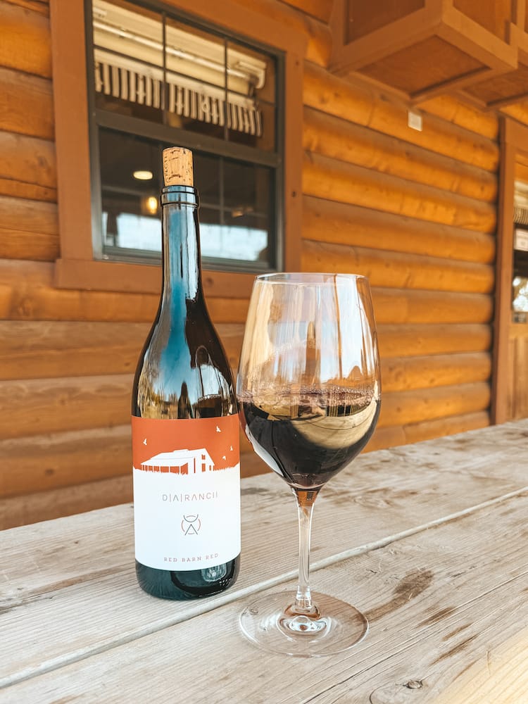A bottle of "Red Barn Red" and a glass of red wine sitting on a wood table at DA Ranch, one of the best wineries in Sedona.