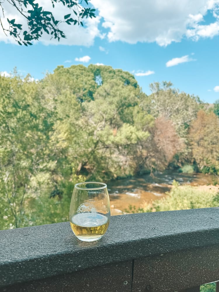 A glass of white wine with a backdrop of the Verde River, lush, green trees. and a blue sky overcast with fluffy white clouds.
