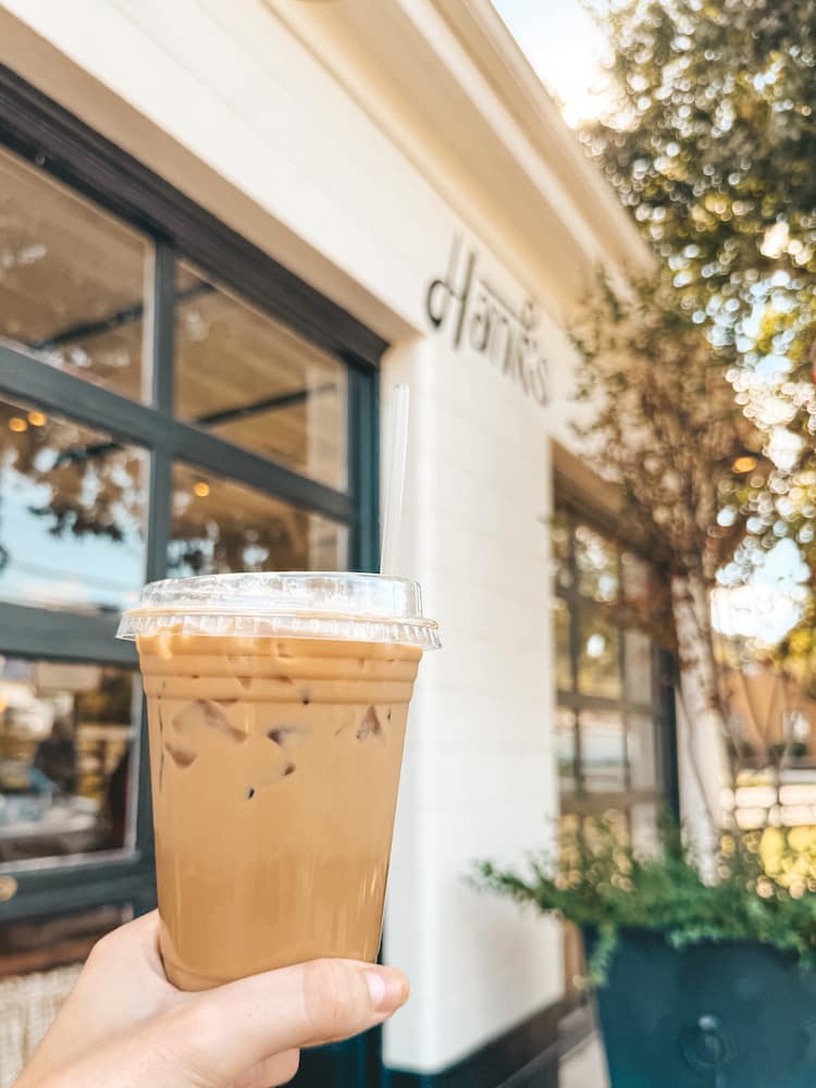 An iced coffee being held in front of the white brick exterior of Hank's Coffee Station, one of the best coffee shops in Franklin