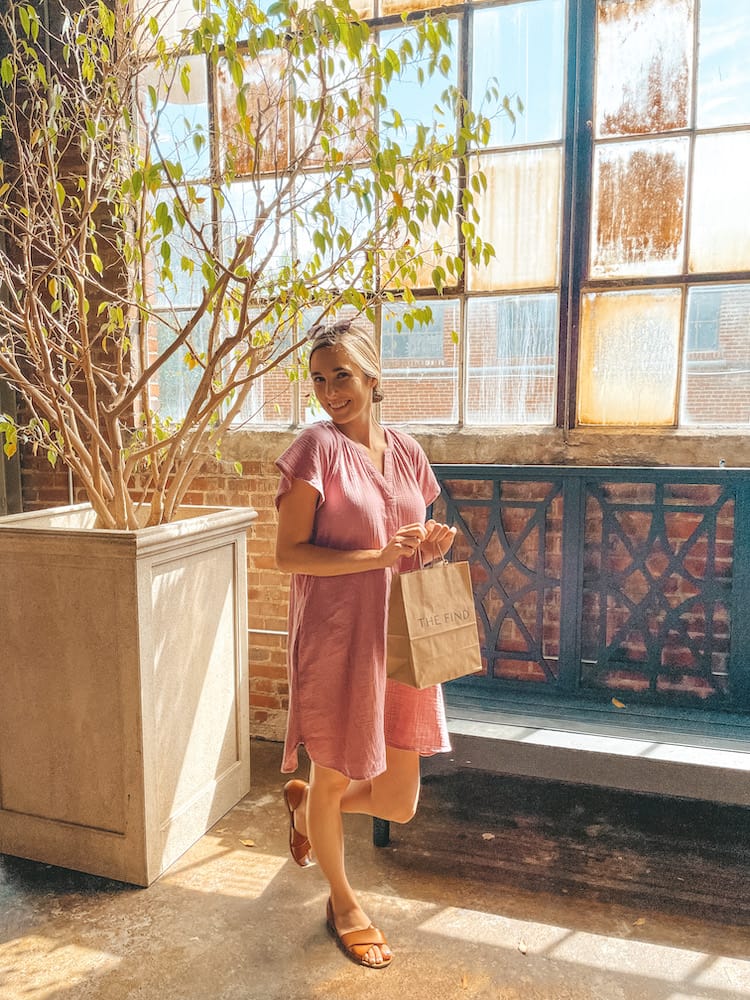 Travel by Brit holding a brown paper and smiling in front of a tree at The Factory at Franklin