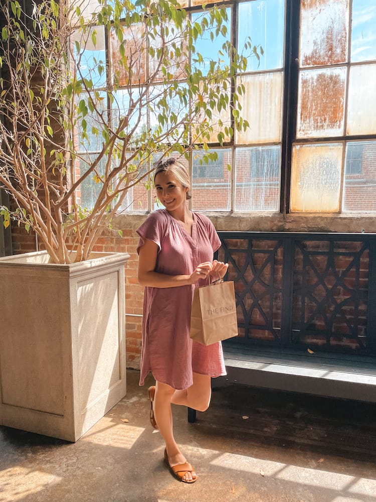 A woman in a pink dress holding a brown paper bag standing in front of a stained glass window inside The Factory at Franklin