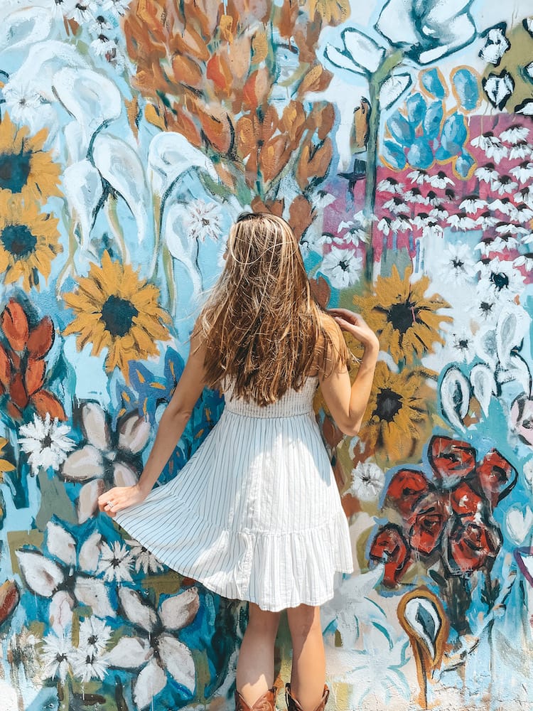 A woman in a white sundress with long brown hair standing in front of a wall painted blue with colorful yellow, pink, orange, red, and white flowers