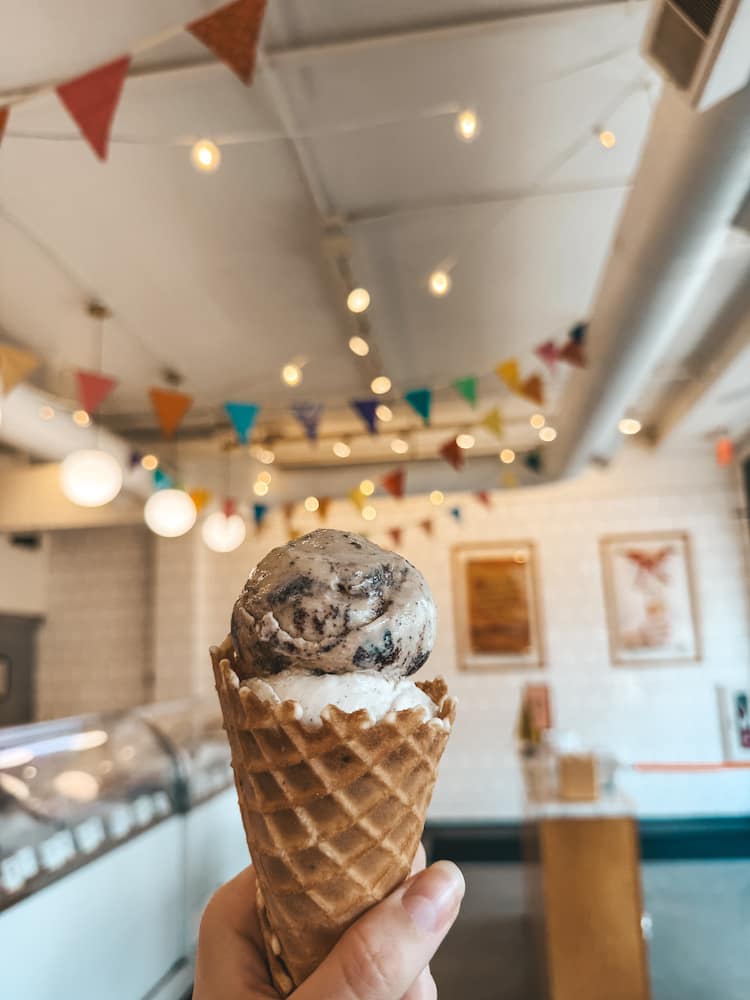 A woman's hand holding an waffle cone ice cream with two scoops in an ice cream shop with several pennant flags and pendant lights hanging from the ceiling
