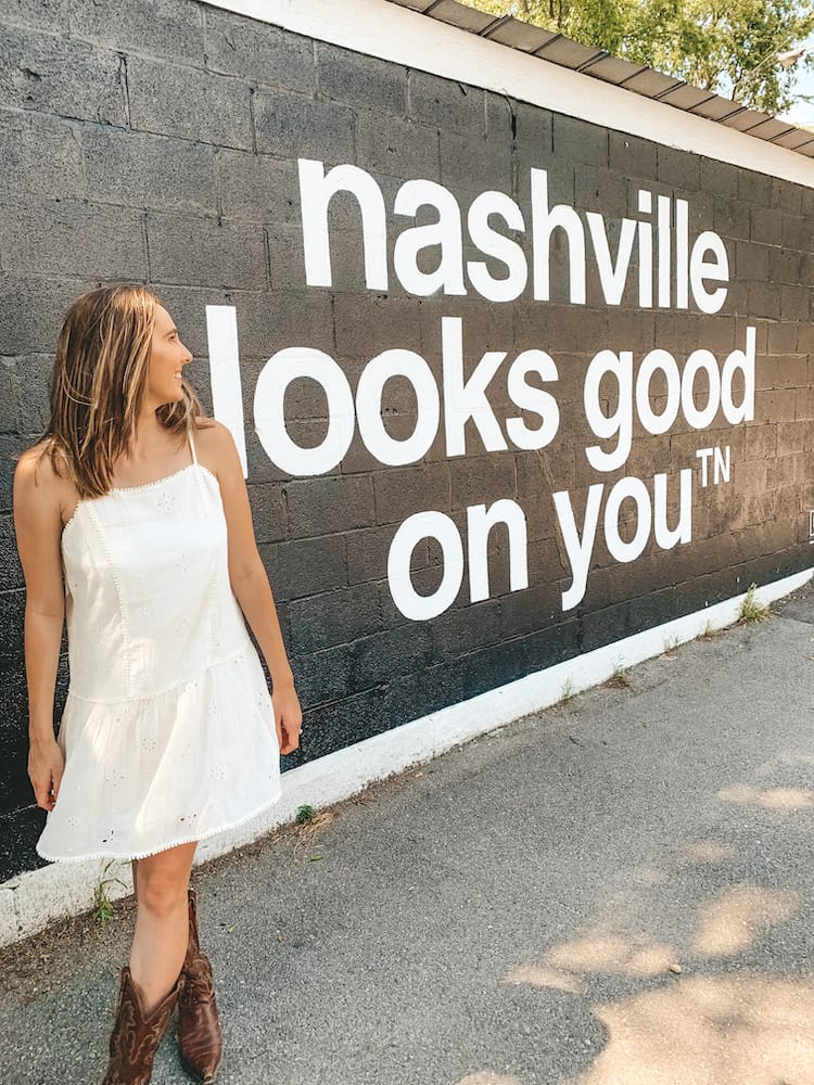 A woman in a white sundress with brown cowboy boots standing in front of a black wall with white lettering that says "Nashville Looks Good on You"