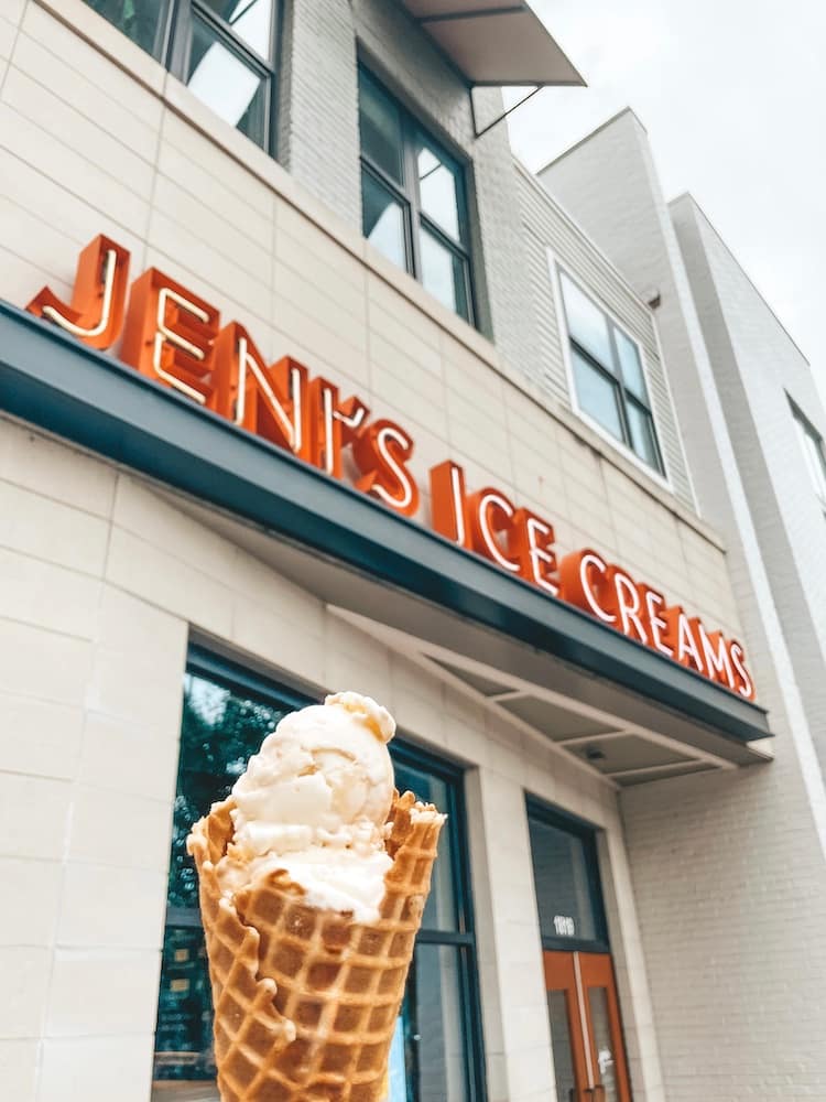 A scoop of ice cream on a waffle cone being held outside the orange neon sign for Jeni's Ice Creams in Nashville