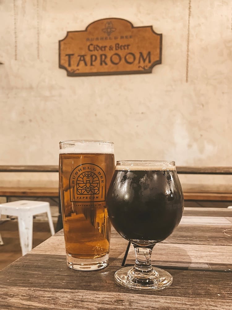 Two beers sitting on a table with the Bushel & Bee Cider & Beer Taproom sign on the wall in the background.