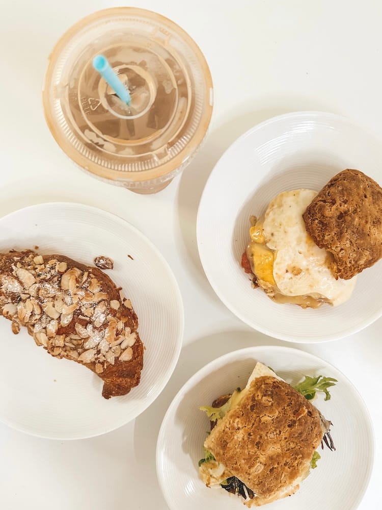 A plate with a croissant, two breakfast sandwiches, and a coffee sitting on a white table.