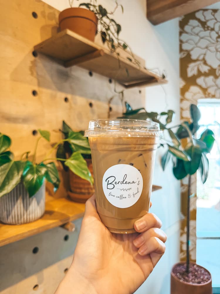 An ice coffee with a white sticker that says "Berdenas" in front of a wall with greenery and yellow and white wallpaper