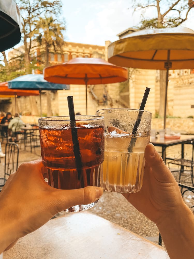 Two people "cheersing" two glasses of red and white sangria in front of colorful red, yellow, and green umbrellas at a Mexican restaurant on the River Walk in San Antonio, Texas