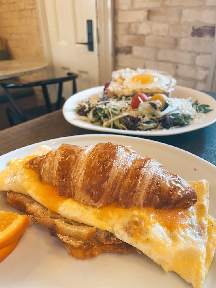 A croissant sandwich with egg and a salad and sunny side up egg sitting on a wooden table with a brick backdrop at one of the best places to eat breakfast during a weekend in San Antonio