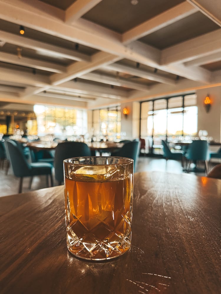 A bourbon cocktail sitting on a brown wooden table with blue chairs and big windows in the background at Fat Ox in Scottsdale.