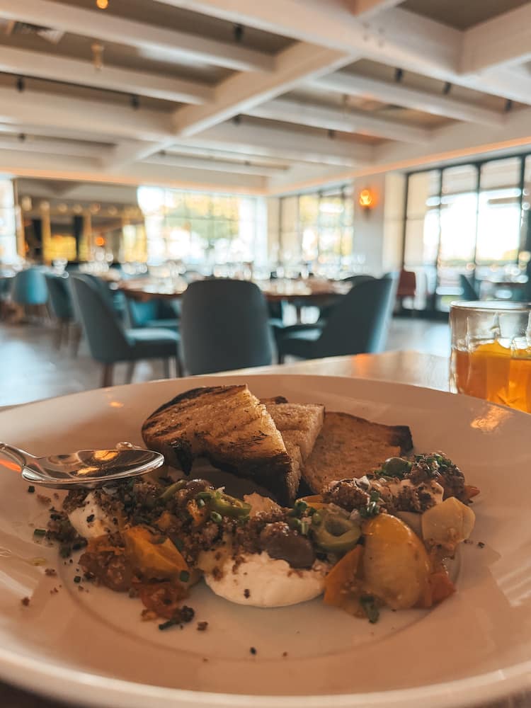 A plate with bread, burrata, olives, and tomatoes sitting on a brown wooden table with blue chairs and big windows in the background at Fat Ox in Scottsdale.